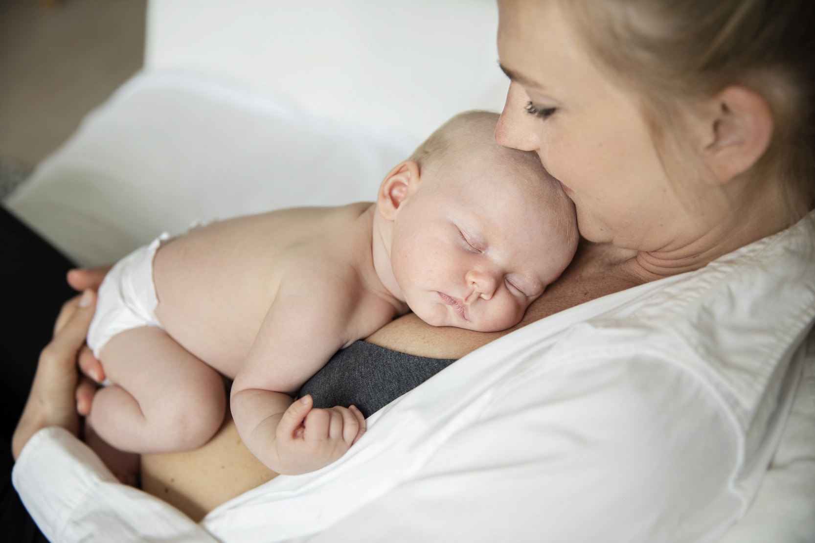 Ett barn i två månaders åldern vilar hud mot hud mot sin mammas bröst med slutna ögon endast iklädd blöja. Mamman har uppknäppt blus.