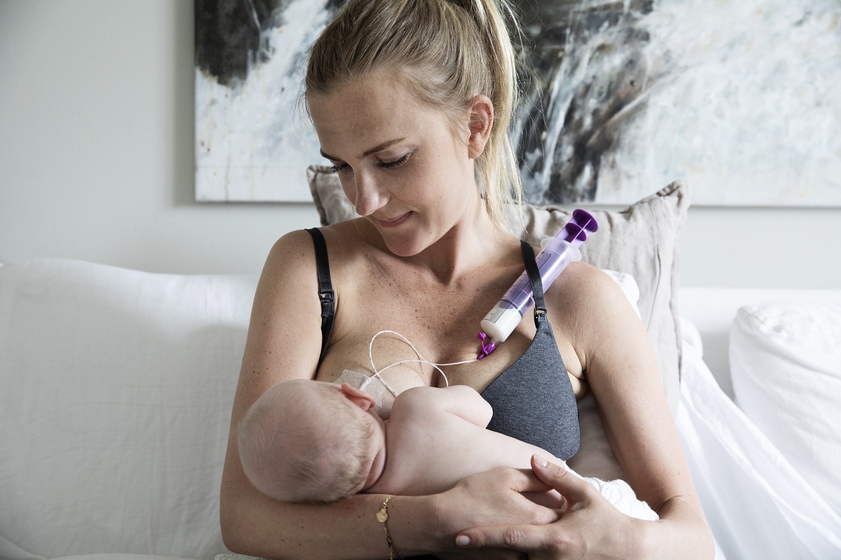 Mamma sitter med bebis i famnen. Bebisen får bröstmjölk genom en sondspruta som är fäst på bröstet.