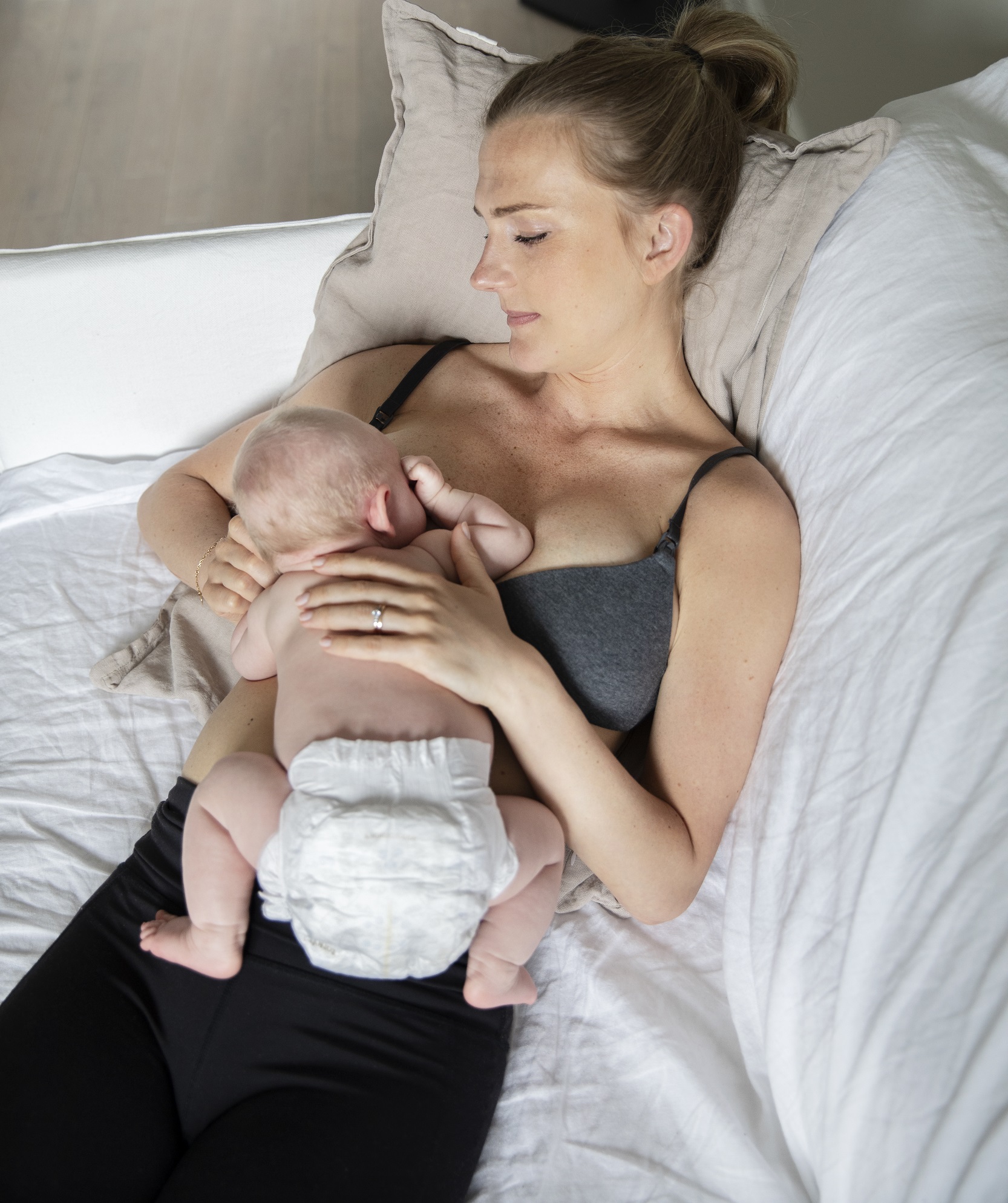 En mamma ligger bekvämt tillbakalutad i en soffa på en kudde i soffhörnet. Hennes barn som är en månad gammal ligger på hennes mage och bröst och ammar vid hennes högra bröst.