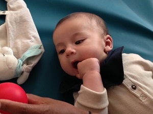 Bebis med fingrar i mun tittar på röd boll. Foto. 