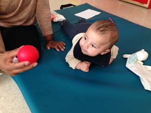Ett spädbarn ligger på en filt på golvet och vrider huvudet för att titta på en räd boll som den vuxna håller i handen.