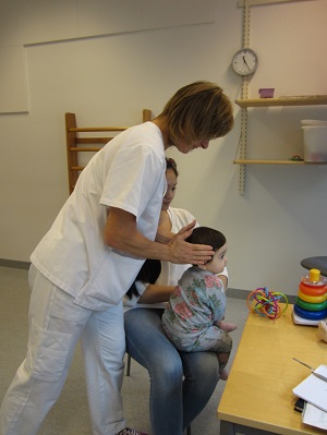 Ett barn i två års åldern sitter i sin förälders knä medans en BHV-sjuksköterska undersöker barnets huvudform.