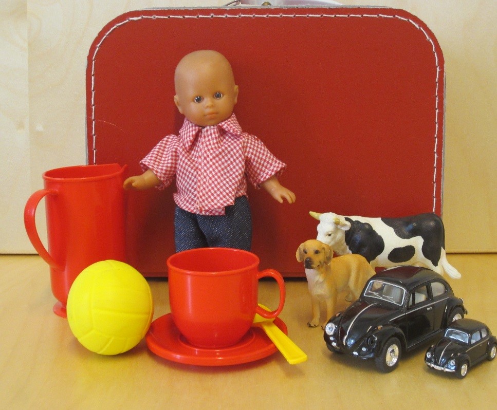 Väska eller låda som innehåller lekföremål: boll, hund, ko, docka, kopp, fat, sked, liten och stor bil. Bild. 