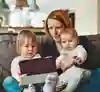 Mamma sitter i soffa med ett sexmånaders barn i knät och ett barn i treårsåldern bredvid. De tittar tillsammans på en surfplatta.